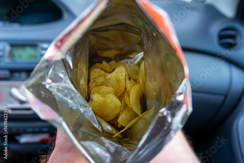 Open bag of chips held inside a car, visible golden snack and part of vehicle dashboard interior.