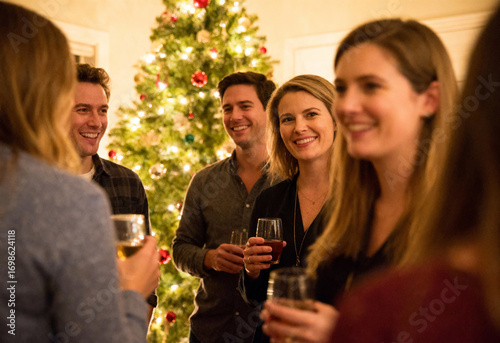 Friends gathering at home for Christmas celebration with young adults smiling and drinking wine near decorated tree in festive evening setting