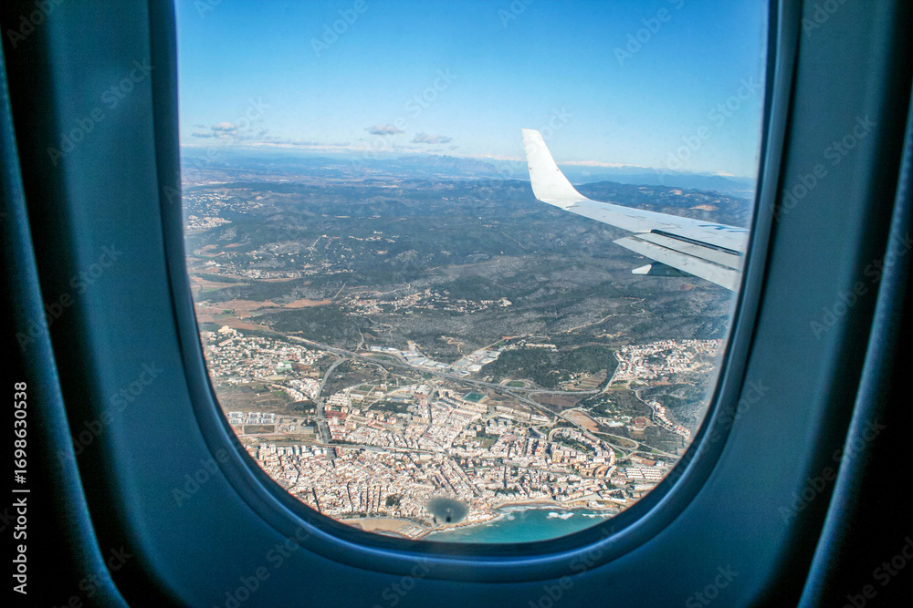 Naklejka premium Aerial view from airplane window: city, coast and mountain landscape under clear blue sky. Travel, vacation, tourism, transportation and adventure concept.