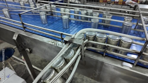 Several metal cans are lined up on a conveyor in a production facility. Workers in the background monitor the machinery's operation, ensuring a smooth flow of the manufacturing process.