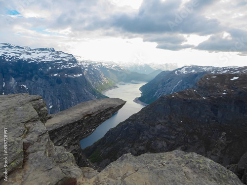 The world-famous Trolltunga rock formation juts out horizontally from a massive cliff, high above the serene blue fjord below, showcasing one of Norway's most spectacular and iconic natural landmarks.