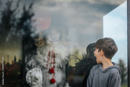A young boy in a gray hoodie stares at a menacing clown figure with bloodstained costume and red balloons inside a glass display, creating a chilling horror-themed scene.