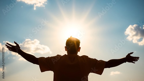 Silhouette of a man with outstretched arms facing the sun against a blue sky with scattered clouds