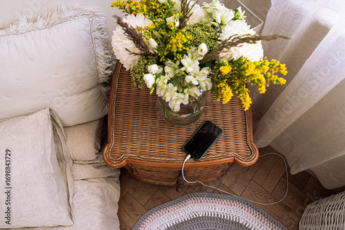 Cozy still life with a smartphone charging on a wicker nightstand decorated with a glass vase of fresh flowers. Aesthetic bedroom interior	