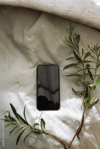 Smartphone with olive tree branches on soft linen bedding, minimal aesthetic flat lay with natural light and earthy tones.	