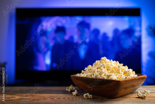 A wooden bowl of popcorn and remote control in the background the TV works. Evening cozy watching a movie or TV series at home