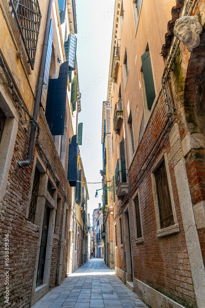 Fototapeta premium Narrow cobblestone street with open shutters in Venice. Charming narrow street in Venice with brick buildings, open wooden shutters, balconies, and soft light illuminating the alleyway.