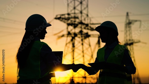 Colleagues women shake hands. Teamwork of power engineers in protective helmets, maintenance of power lines in outdoors. Two construction engineers work together on an electrical transmission line.