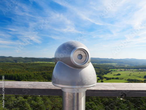 A modern metallic viewing telescope stands on a wooden platform overlooking a vast green valley and distant forested hills under a clear blue sky