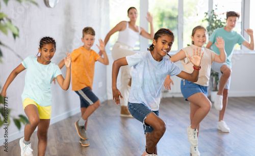 Positive African male child dances vogue in choreographic school. Group of young people in sportswear trains together with classmates in gym before competitions, teacher take part in class background