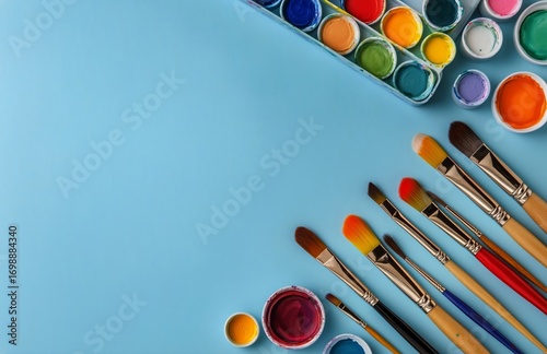 Watercolor Paints and Brushes Surrounding Blank White Paper on Blue Background.