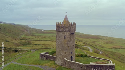 Aerial orbit of Doonagore Castle, showcasing its imposing tower, lush green coastline, and the Atlantic Ocean in the background.