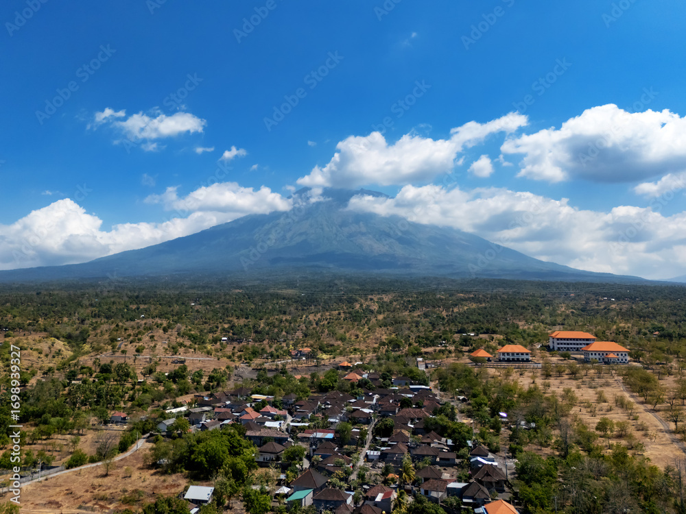 Φωτογραφία Aerial view of Mount Agung Bali with clear blue sky, fluffy clouds, and traditional village houses in the foreground surrounded by green and dry landscapes