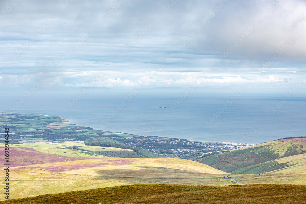 Fototapeta premium Laxey, Isle of Man 08,05,2023 Beautiful view of the ocean and a town in the distance. The sky is cloudy and the water is calm Isle of Man -