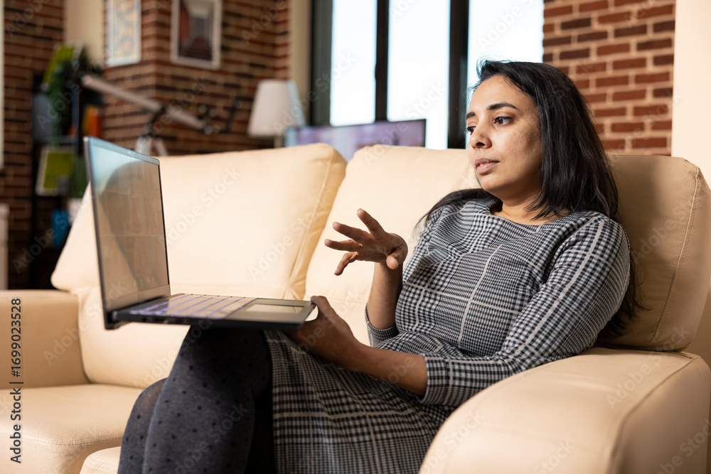 Obraz premium Indian female executive working from home, speaks towards laptop, addressing company issues through video call. Focused woman on sofa, virtually communicating with remote colleagues.