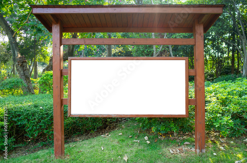 Wooden outdoor signboard with blank white display panel under a roofed structure, placed in a green public park. Mockup copy space for text, information, community announcement or advertisement.