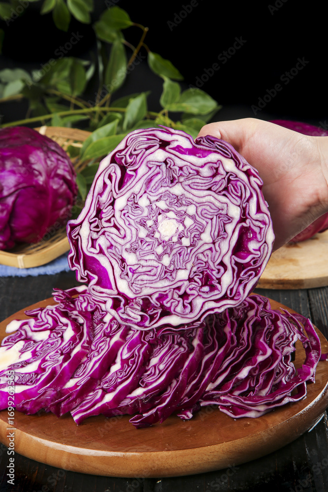 Obraz premium Hand Slicing Fresh Purple Cabbage on Wooden Cutting Board Showing Vibrant Cross-Section and Leafy Texture