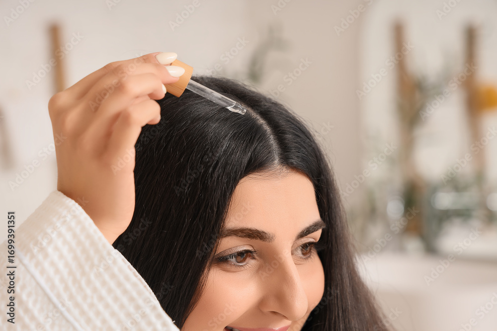 Naklejka premium Young brunette woman applying serum for hair growth in bathroom, closeup
