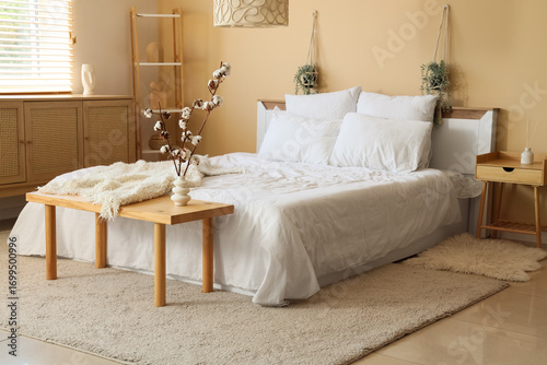 Vase with cotton flowers on wooden bench near beige wall in interior of bedroom
