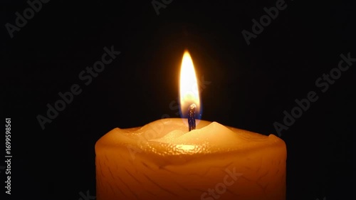 Close up macro shot of a single lit candle flame flickering gently in darkness with a warm orange glow illuminating the wax surface and creating a