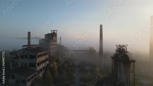 Drone footage capturing an aerial view of an abandoned industrial factory shrouded in morning mist at dawn, urban decay concept.