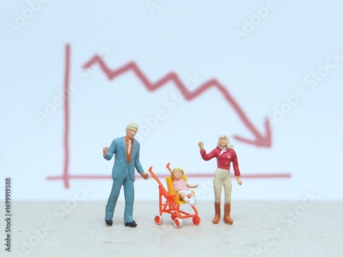 Mini toy of action figure at table with blurred background. Miniature photography concept design. Minifigure of father, baby at stroller and mother character with down arrow chart.