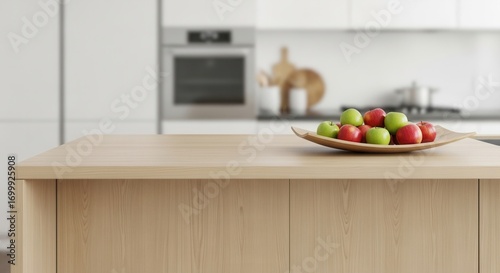 A light wooden island counter holds a leaf-shaped bowl of red and green apples A blurred kitchen background shows white cabinets and a built-in oven