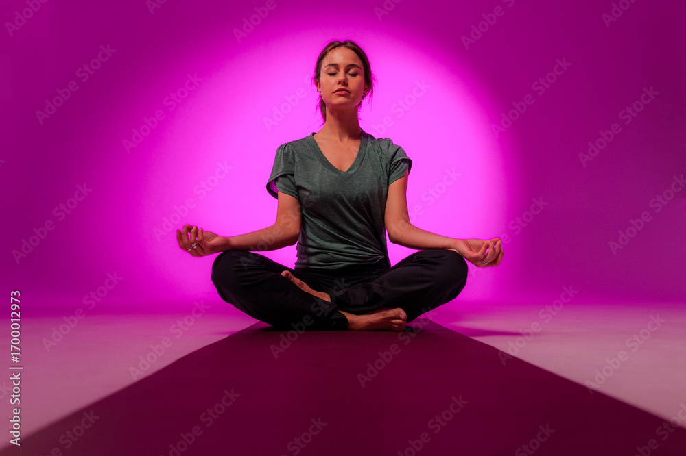 Fototapeta premium Young woman meditating in lotus pose on yoga mat with purple background