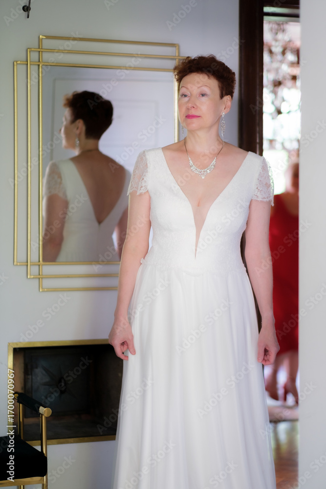 Fototapeta premium A woman in a white dress is standing. There is a mirror behind the woman. The woman's reflection in the mirror is visible.