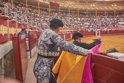 Two matadors in traditional embroidered suits stand by the arena wall with pink and yellow capes during a bullfight in Almeria, Spain. Tradition, culture