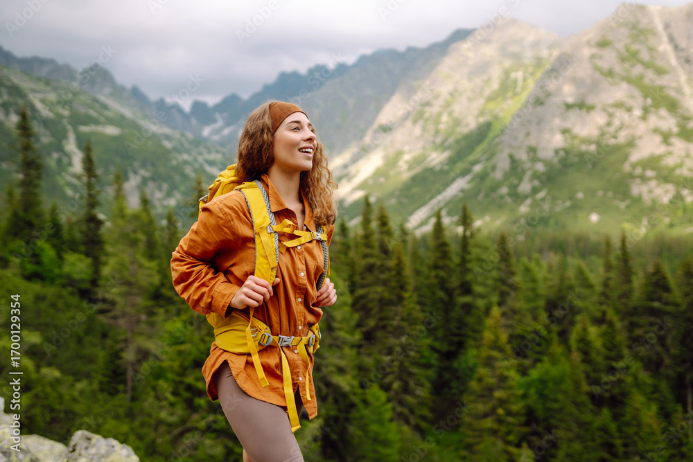 Naklejka premium A female hiker with a yellow backpack on a rocky path on a sunny day. Happy woman enjoying the mountain landscape and feeling freedom in the open air. Nature, adventure concept.