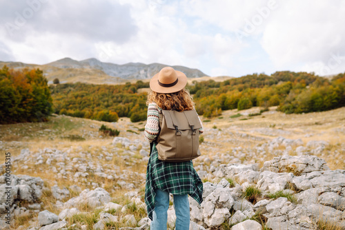A rear view of a female hiker with a backpack and a hat walking along a rocky trail at sunset. Young woman enjoying the mountain scenery, admiring the views. Adventure, freedom and weekend concept.