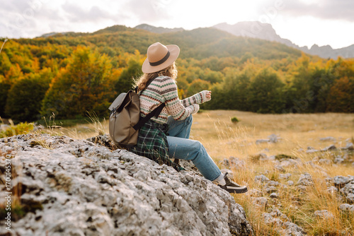 A rear view of a female hiker with a backpack and a hat walking along a rocky trail at sunset. Young woman enjoying the mountain scenery, admiring the views. Adventure, freedom and weekend concept.