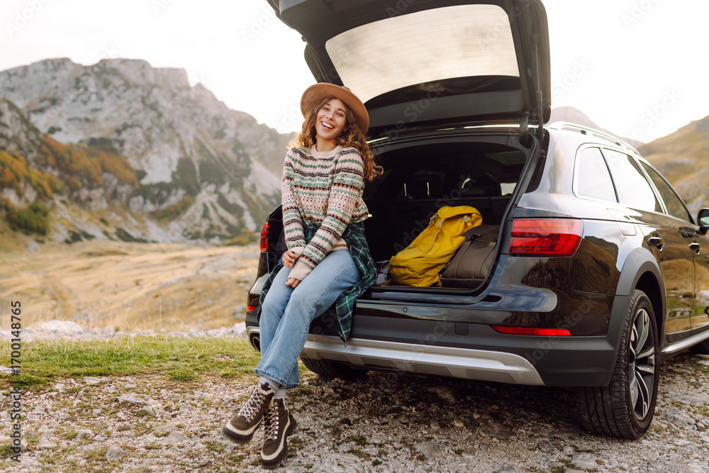 Naklejka premium A joyful woman in a hat sits in the trunk of a car and admires the mountain landscape. A woman on a car trip enjoys the sunset and mountains. Transport concept, adventure.