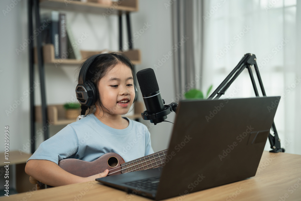 Fototapeta premium Young girl with headphones sings and plays ukulele in front of laptop during online music lesson in modern home setup with natural light.