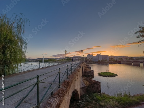 Pedestrian bridge over the Tua River in Mirandela, Portugal, leads to the city center at sunset. Evening light, calm water, and urban skyline create a tranquil and scenic connection.