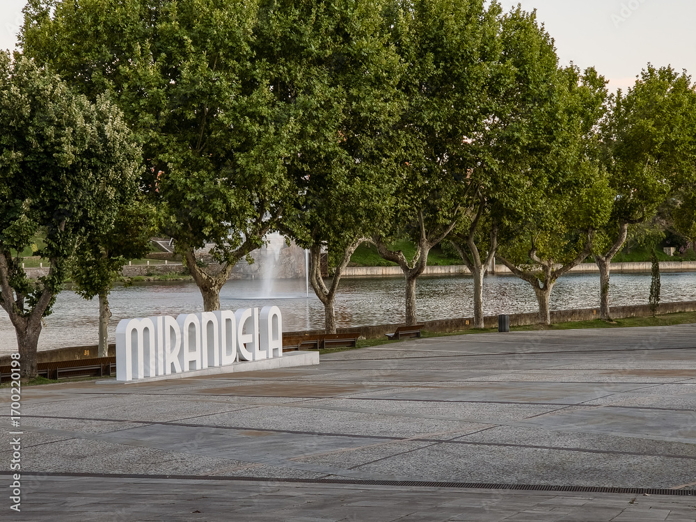 Naklejka premium Mirandela, Portugal: The riverside square with large trees and the Tua River creates a welcoming urban space, where the iconic city sign invites residents and visitors to enjoy leisure and nature.