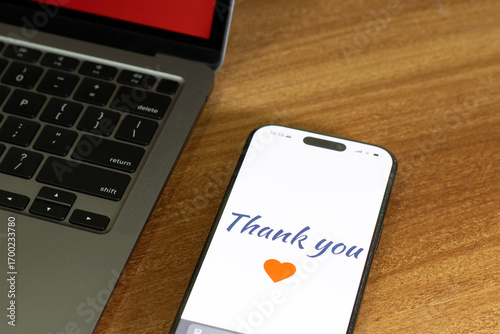 Mobile screen with Thank you message with heart symbol on the wooden table along with the laptop.