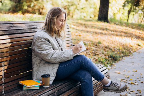 Young woman writing in notebook on bench in autumn park.