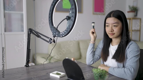Young Thai woman recording a beauty video vlog at home. She talks to the camera with confidence while applying makeup and sharing skincare tips.
