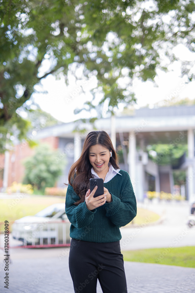 Fototapeta premium Asian female student in warm clothes using phone to chat with friends and ready for video call while in university.
