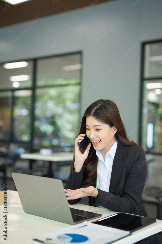 Fototapeta premium A beautiful businesswoman in a suit is looking at a computer screen while talking on her mobile phone. She is sitting in an office and using modern technology.