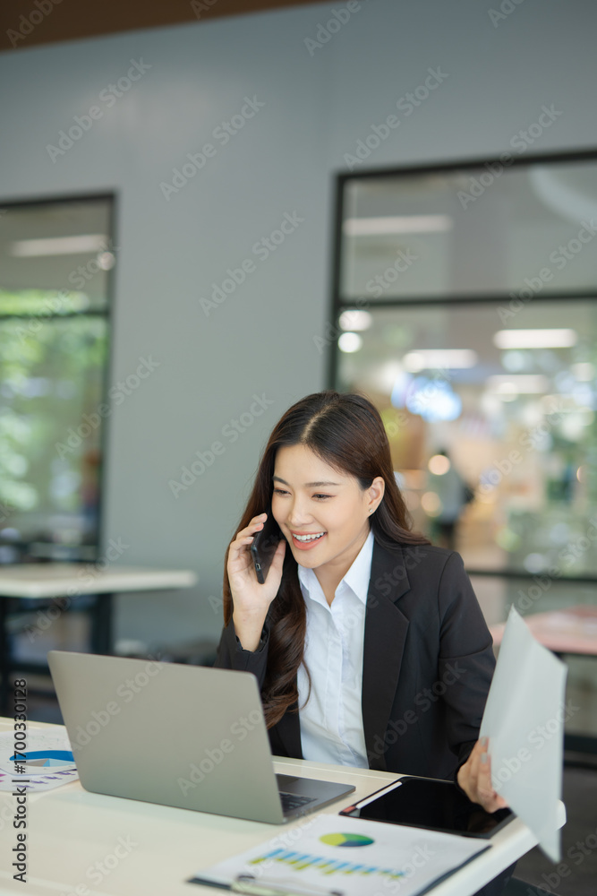Fototapeta premium Asian businesswoman working at desk using laptop and notebook, talking with client or modern office building.
