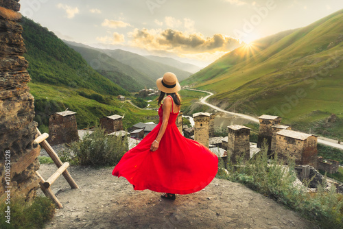 Woman in red dress looking at medieval towers of Ushguli Georgia during sunset with mountains and Enguri valley landscape