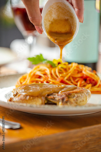 A gourmet dish with roasted chicken breast topped with sauce, served alongside spaghetti pasta with tomato sauce, photographed with sauce being poured over the chicken.