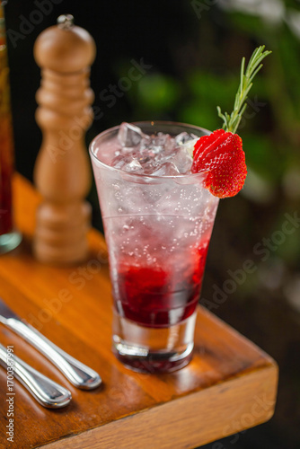 A refreshing strawberry cocktail served in a tall glass with ice, layered with red syrup, and garnished with a fresh strawberry slice and a rosemary sprig.