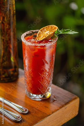 A classic Bloody Mary cocktail served in a crystal glass with salted rim, ice cubes, and garnished with a dried lime slice and fresh parsley, set on a wooden table.