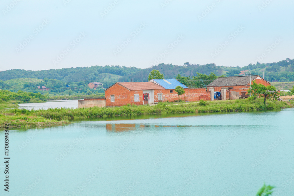 Obraz premium Rural brick buildings with solar panels reflected in tranquil lake water surrounded by green hills and natural countryside landscape