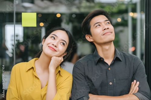 Happy Asian man and woman looking up with positive expressions in office. Creative team standing in front of glass wall with sticky note, concept of inspiration, teamwork and planning.