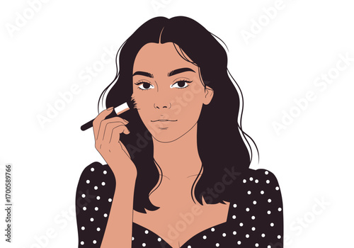 A woman with dark hair and a dotted dress applies makeup with a brush on her cheek